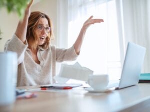 Woman in glasses extremely surprised with new higher taxes watching, throwing bills at laptop in home office. Small business troubles, bull market or money savings concept image